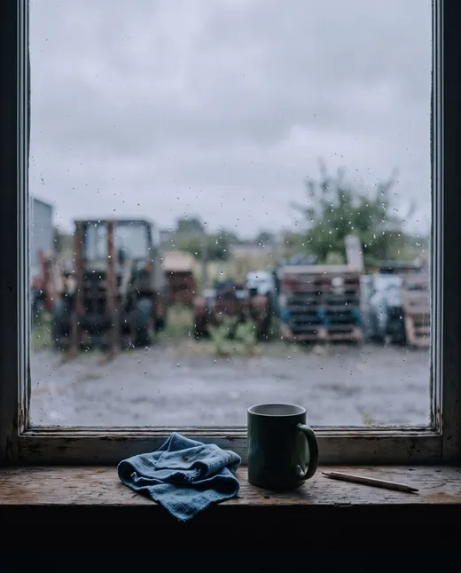 Rain on a workshop window, a mug and a folded rag on the sill, the yard grey and out of focus beyond.
