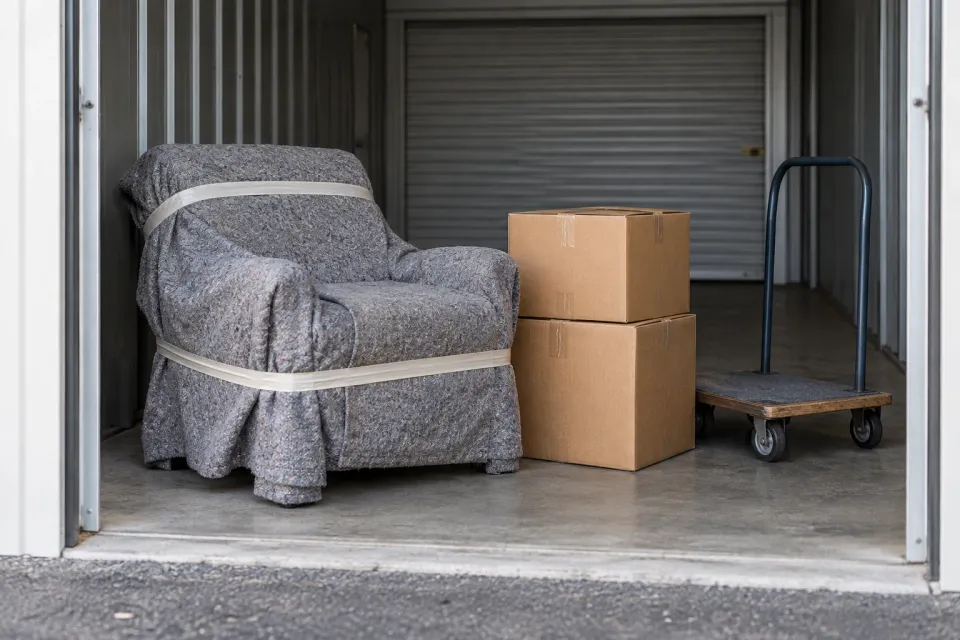 Editorial illustration: A blanket-wrapped armchair, two sealed cartons and a trolley at a storage entrance.